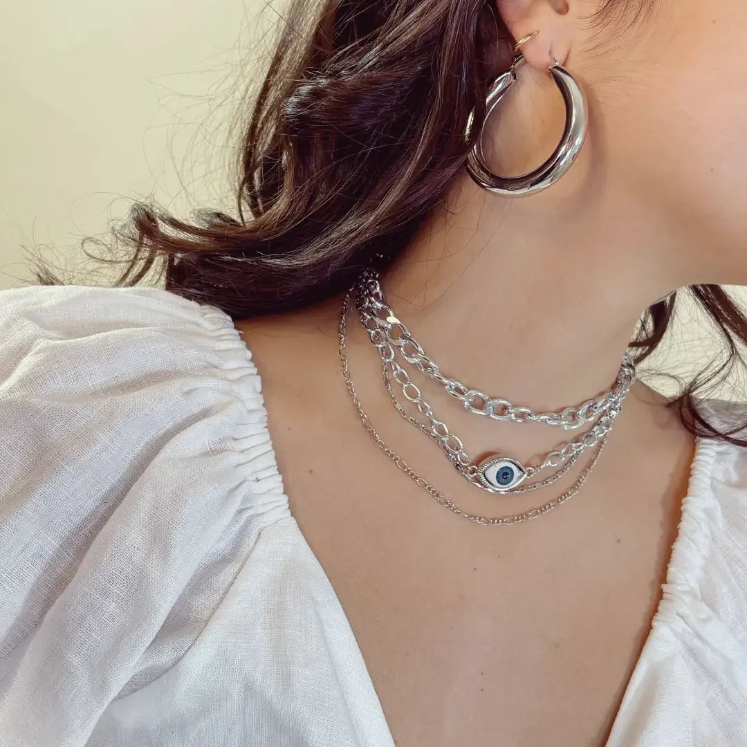 Woman wearing silver chunky hoop earrings and layered silver chain necklaces with an eye pendant, white ruched sleeve top