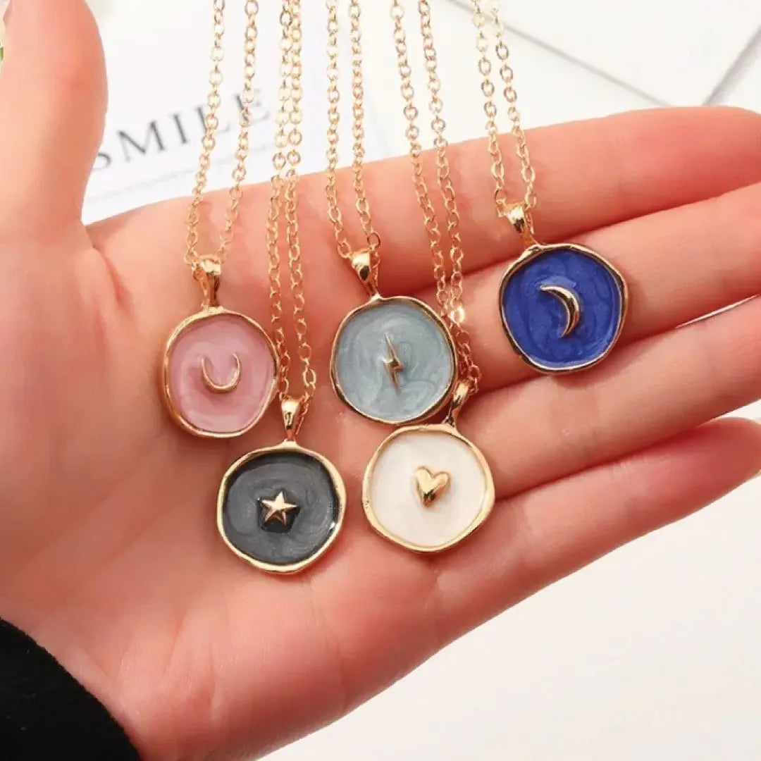 Hand holding five gold chain necklaces with round pendants in pink, gray, blue, black, and white featuring crescent moon, lightning bolt, star, and heart designs