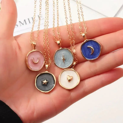 Hand holding five gold chain necklaces with round pendants in pink, gray, blue, black, and white featuring crescent moon, lightning bolt, star, and heart designs
