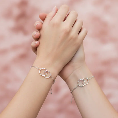 Two hands clasped wearing silver friendship bracelets with interlocking circles on pink background