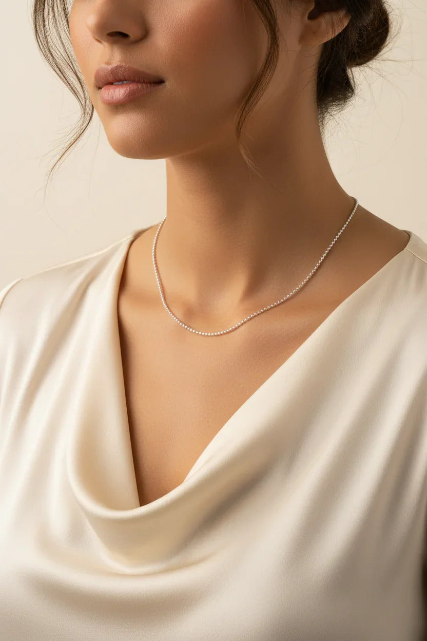 Close-up of a woman wearing a delicate silver chain necklace with a cream satin draped top