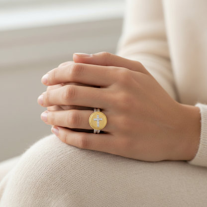 Close-up of a hand wearing a gold round ring with a diamond cross design on a cozy ivory fabric background