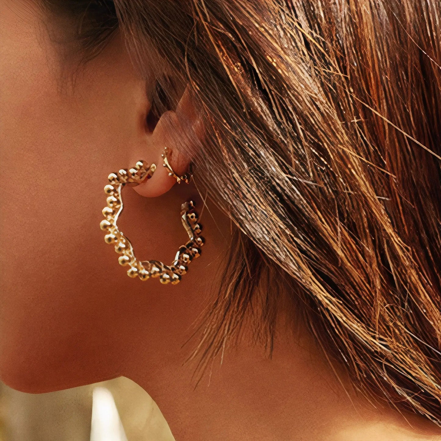 Close-up of a woman's ear wearing textured gold hoop and small gold cuff earrings, highlighting luxury jewelry details