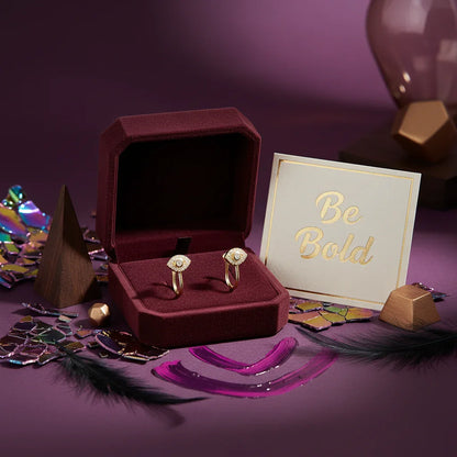 Gold eye-shaped ear cuffs with diamonds in a burgundy velvet box beside a 'Be Bold' gold-lettered card on a purple surface