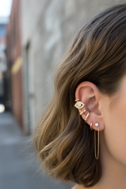 Close-up of a woman's ear adorned with gold ear cuffs, including a diamond-studded evil eye design and chain drop earring