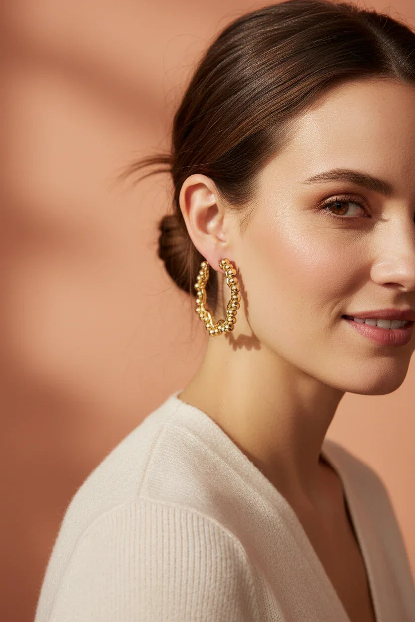 Close-up of a woman wearing textured gold hoop earrings with a casual cream top and soft peach background