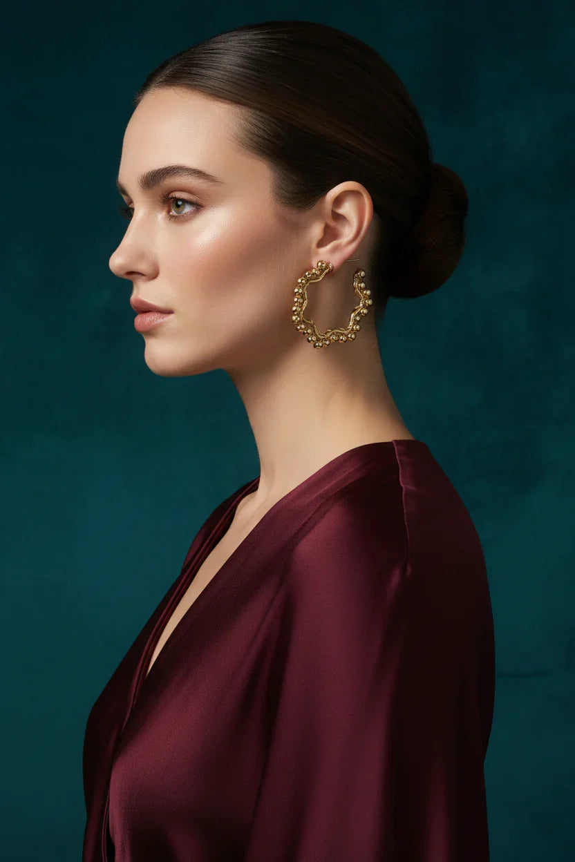 Profile of woman with sleek bun wearing textured gold hoop earrings and deep burgundy satin dress against dark teal background
