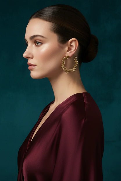 Profile of woman with sleek bun wearing textured gold hoop earrings and deep burgundy satin dress against dark teal background