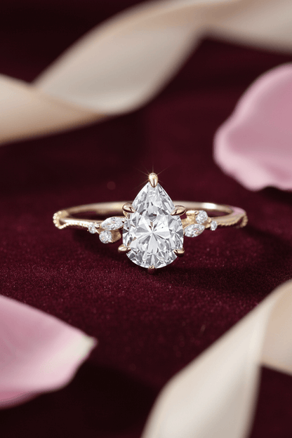 Pear-shaped diamond ring with gold band and small side diamonds on burgundy velvet with pink petals