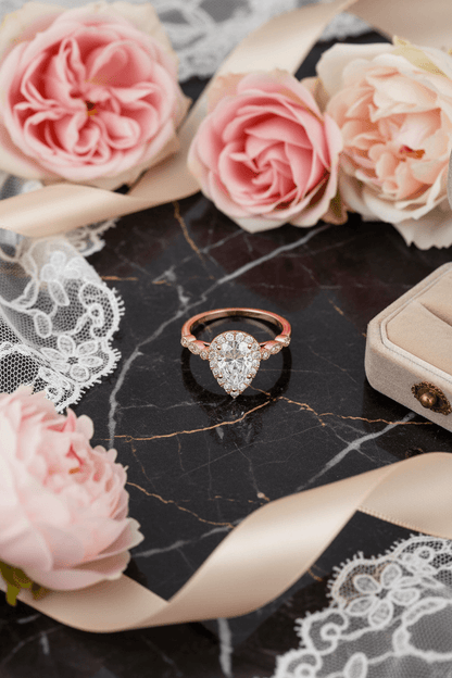 Elegant pear-shaped diamond engagement ring with rose gold band surrounded by pink roses and lace on black marble