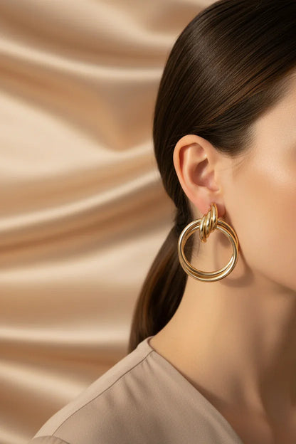 Close-up of a woman wearing large gold statement hoop earrings against a smooth champagne fabric background