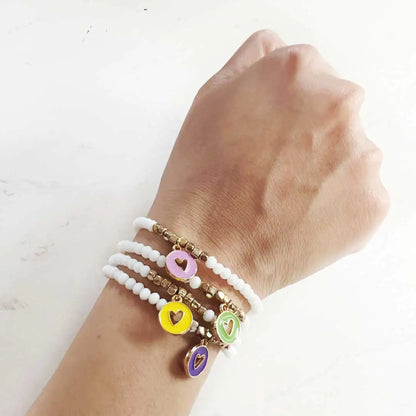 Wrist wearing layered white and gold bead bracelets with colorful heart charm pendants in pink, yellow, green, and purple