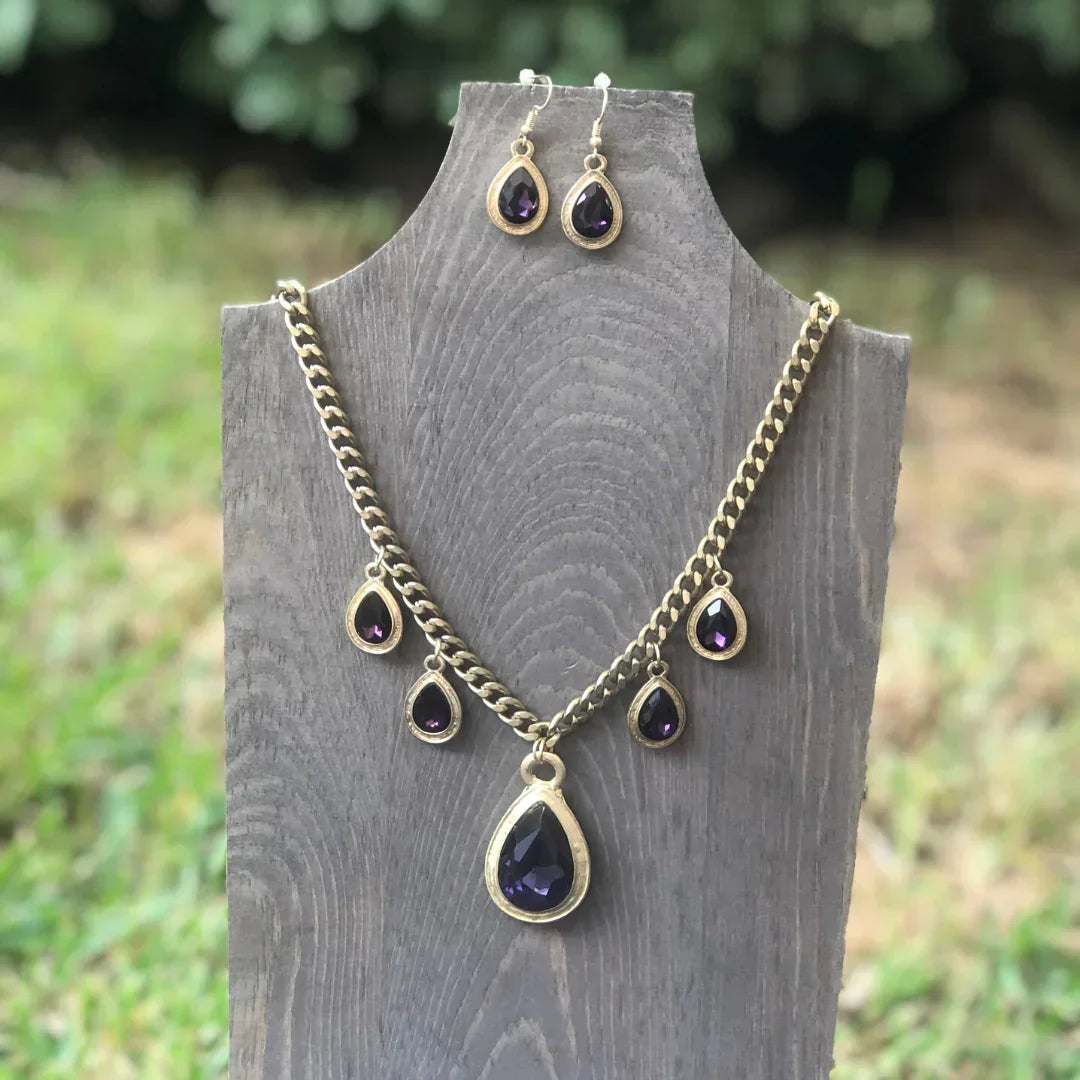 Gold-tone necklace and earrings set with teardrop-shaped dark purple gemstones on wooden display