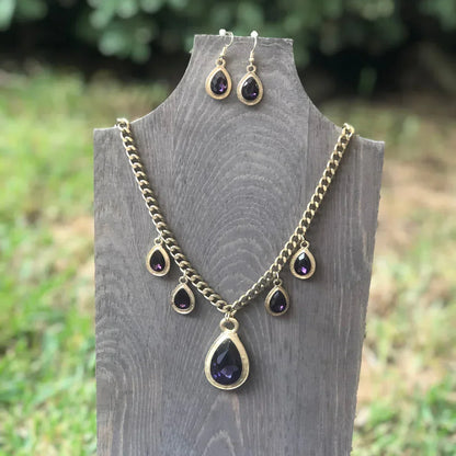 Gold-tone necklace and earrings set with teardrop-shaped dark purple gemstones on wooden display