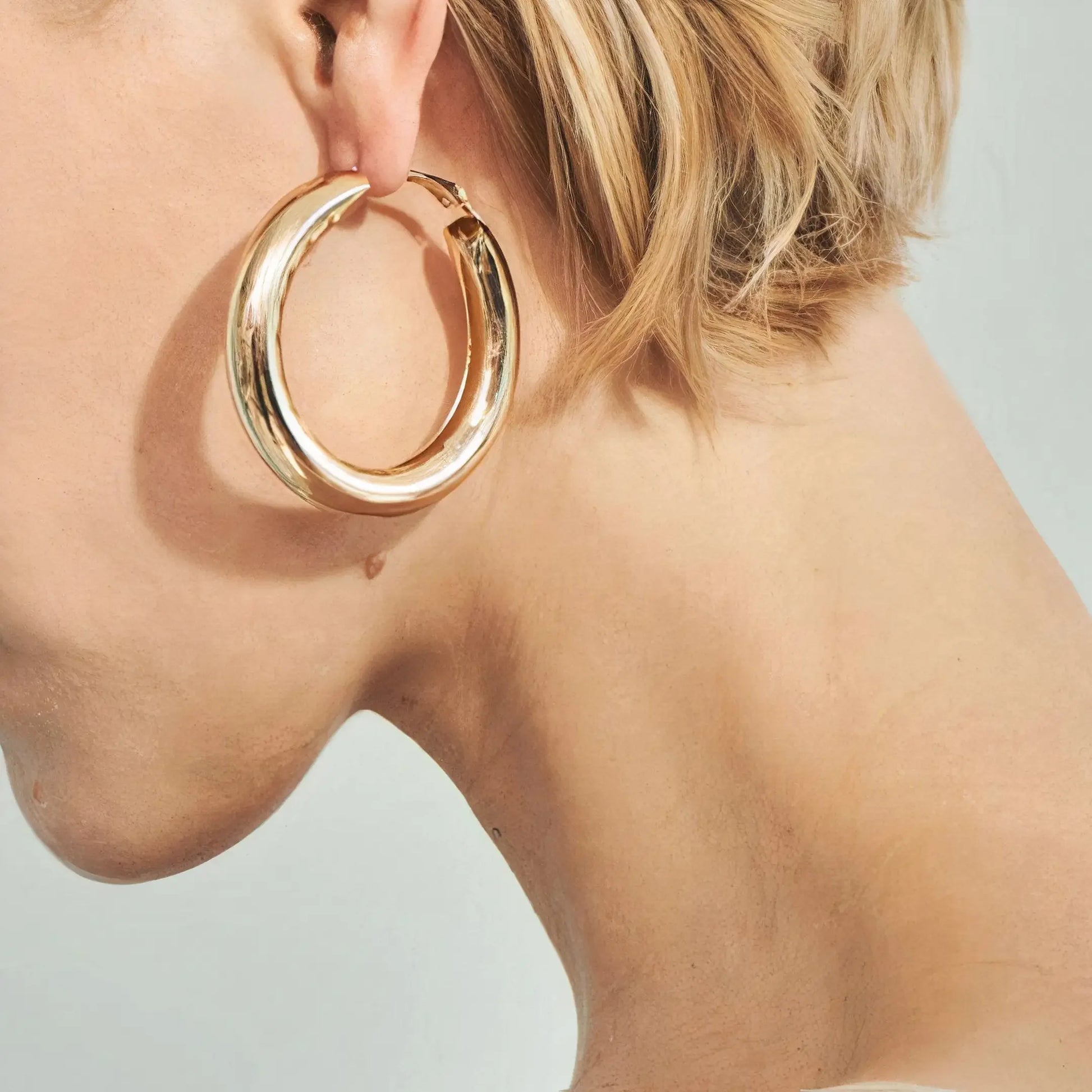 Close-up of a woman's ear wearing a large polished gold hoop earring against a light background