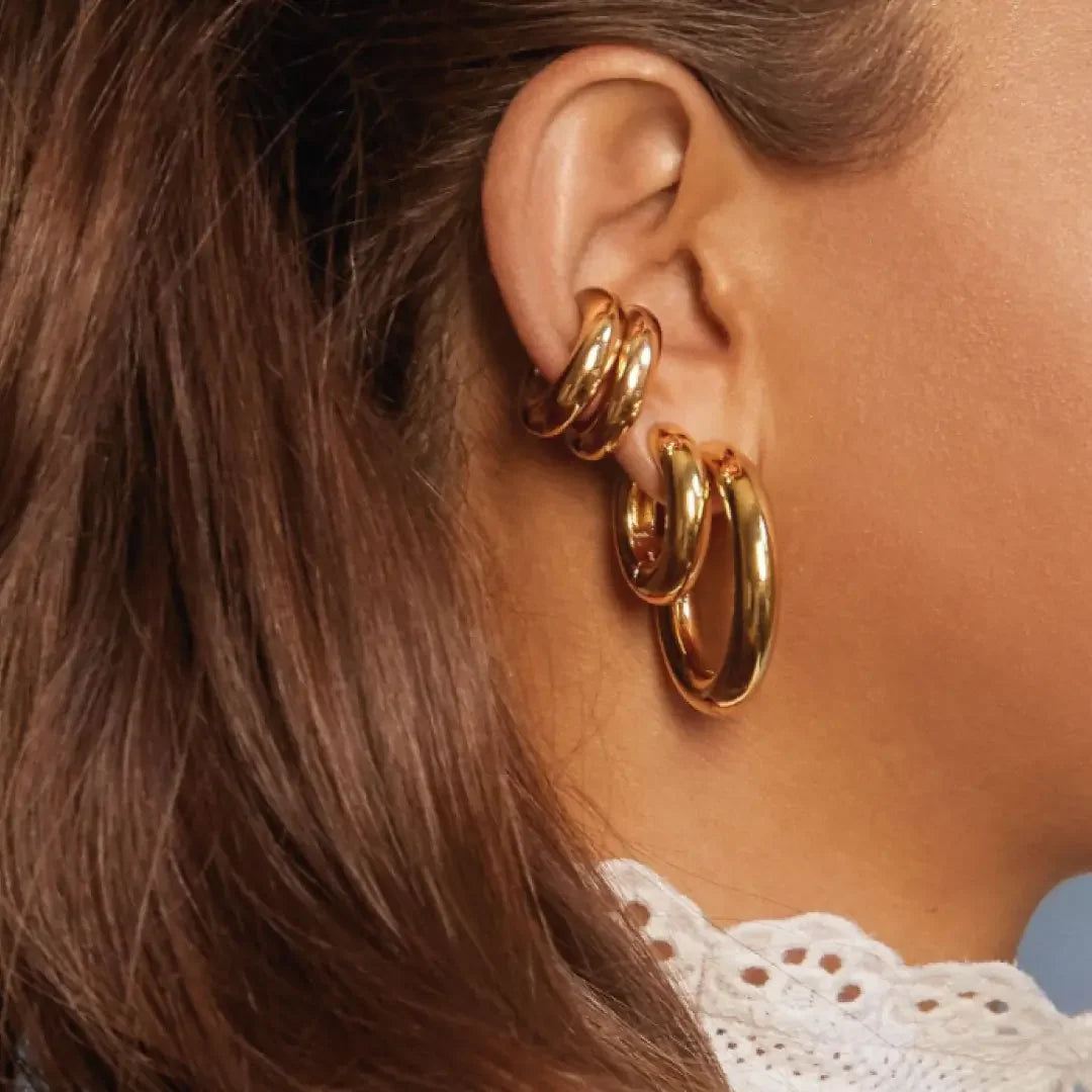 Close-up of a woman's ear adorned with three polished gold hoop earrings, styled with brown hair and white lace collar