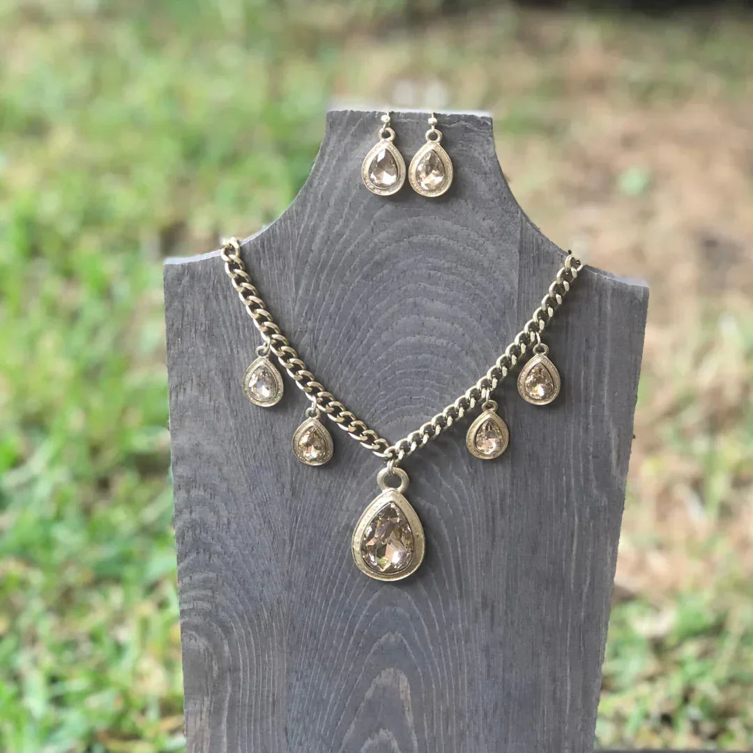 Gold-tone necklace and matching teardrop earrings with clear gemstones displayed on a wooden stand outdoors