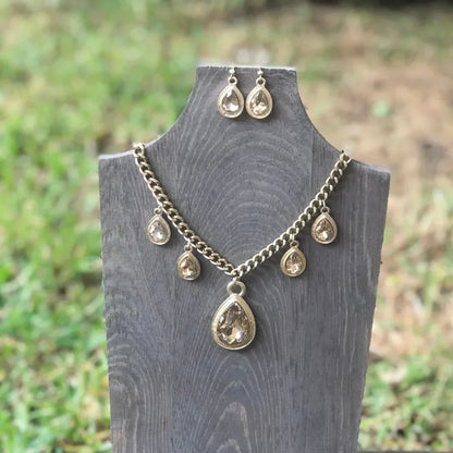Gold-tone necklace and matching teardrop earrings with clear gemstones displayed on a wooden stand outdoors